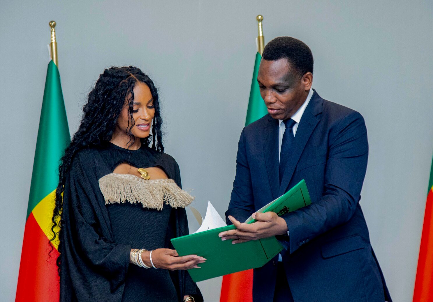 Singer Ciara smiling and holding her Benin passport after being granted citizenship, surrounded by a Beninese official during an official ceremony.