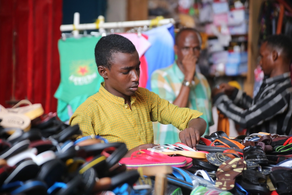 A young Kenyan vendor looks at his phone after receiving a small Hustler Fund loan, surrounded by a struggling market stall
